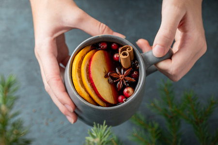 Hand holding a cup of red mulled wine with spices, orange, apple slices and cranberry on a dark...