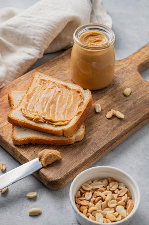 Peanut butter toast on a wooden board on a light background with scattered nuts, napkin and knife. Concept for a sweet vegetarian diet and healthy snacks for an American breakfast.の写真素材