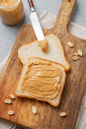 Peanut butter toast on a wooden board on a light background with scattered nuts and knife. Concept for a sweet vegetarian diet and healthy snacks for an American breakfast. Top view, copy space.の写真素材
