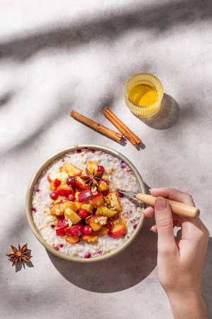Hand holding spoon in to plate of oatmeal with apple, cranberry and cinnamon on a light background with honey and morning shadows. Concept for a healthy vegan meal for breakfast. Top view, copy space.の写真素材