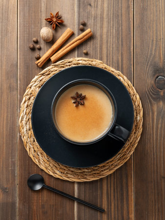 Traditional Indian masala chai tea with milk and spices in a black cup on a dark wooden background with cinnamon, anise and spoon. A cozy hot drink concept for a menu or banner for an ethnic cafe or restaurant. Top view, copy space.