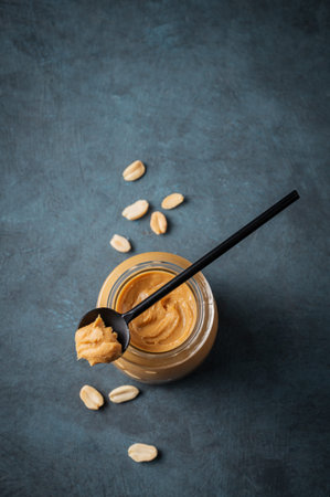 Creamy peanut butter in a jar on a dark background with roasted nuts and spoon close up. Concept for...