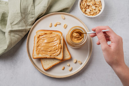 A hand holds a spoon with creamy peanut butter in a jar on a plate with toast and roasted nuts on a...