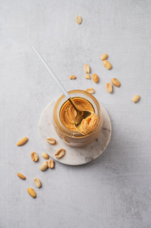 Creamy peanut butter in a jar on a light background with scattered roasted nuts and spoon close up. Concept for a healthy, nutritious vegetarian snack for an American breakfast. Top view, copy space.の写真素材