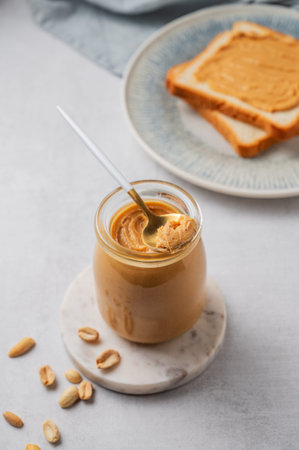 Creamy peanut butter in a jar on a light background with toasted bread, roasted nuts, spoon and napkin close up. Concept for a natural, nutritious vegetarian product. Top view, copy space.の写真素材