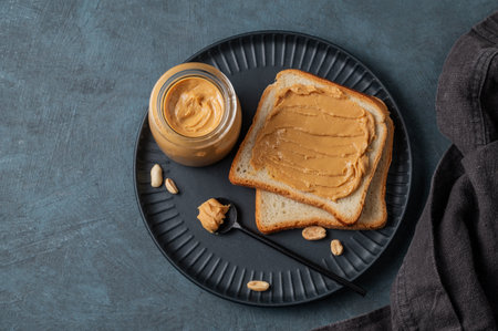Peanut butter toast on a black plate on a dark background with nuts, napkin and spoon. Concept of a nutritious vegetarian spread and healthy snack for an American breakfast. Top view, copy space.の写真素材
