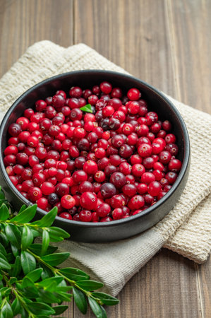 Fresh cranberries in a dark bowl on a wooden background with green branch and napkin. Concept of healthy natural wild berries in rustic style. Top view, copy space.の写真素材