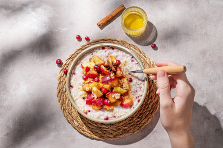 Hand holding spoon in to plate of oatmeal with apple, cranberry and cinnamon on a light background with honey and morning shadows. Concept for a healthy vegan meal for breakfast. Advertising menu or banner cafe. Top view, copy space.の写真素材