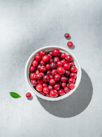 Fresh cranberries in a white bowl on a light background with green leaf and sunlight. Concept of...