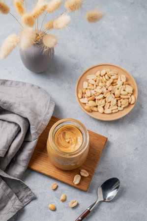 Creamy peanut butter in a jar on a wooden board on a light background with a scattering of roasted nuts, a spoon, and a napkin. Concept for a natural, nutritious vegetarian product. Top view, copy space.の写真素材