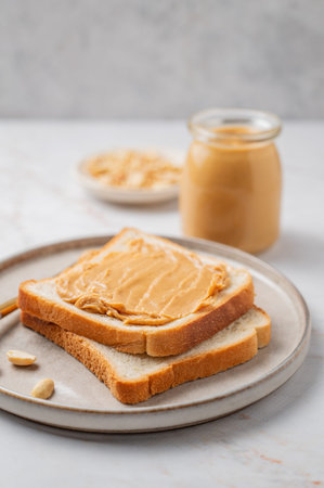 Peanut butter toast on a plate on a light marble background with nuts and knife close up. Concept of...