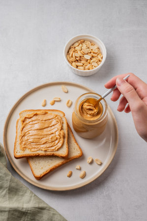 A hand holds a spoon with creamy peanut butter in a jar on a plate with toast and roasted nuts on a light background. Concept for a healthy, nutritious vegetarian snack for an American breakfast. Top view, copy space.の写真素材