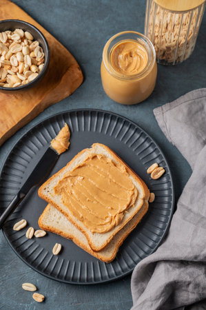 Peanut butter toast on a black plate on a dark background with nuts, napkin and knife. Concept of a...