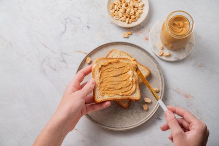 A hand spreads peanut butter on toast over a plate on a light marble background with nuts and knife. Concept for a healthy, nutritious vegetarian snack for an American breakfast. View from above.の写真素材