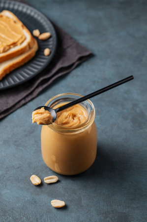 Creamy peanut butter in a jar on a dark background with toasted bread, nuts, spoon and napkin....