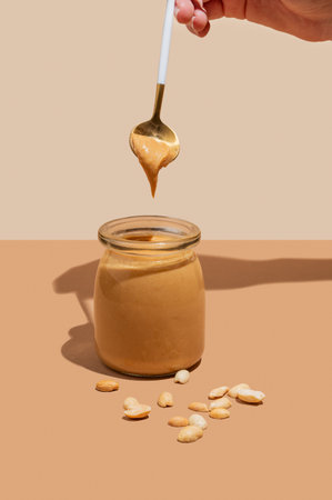 A hand holds a spoon with peanut butter over a glass jar on a beige background with scattered nuts and shadow. Concept for a tasty and healthy snack for sweet breakfast sandwiches. Front view, copy space.の写真素材