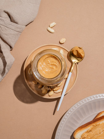 Peanut butter in a glass jar and spoon on a beige background with toast, scattered nuts and napkin. Concept for a delicious and healthy snack for sweet breakfast sandwiches. Top view, copy space.の写真素材