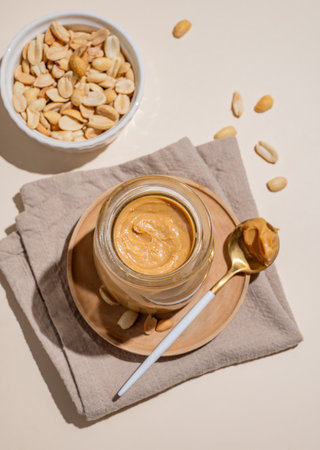 Peanut butter in a glass jar on a napkin on a light background with scattered nuts, spoon and shadow. Concept for a delicious and healthy snack for sweet breakfast sandwiches. Top view, copy space.の写真素材