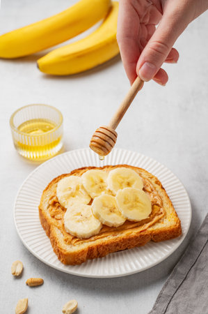 A hand holds a honey spoon over toast with peanut butter and banana on a white plate on a light background with fruits. Concept of nutritious and healthy snacks for breakfast.の写真素材