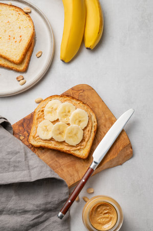 Peanut butter toast and banana on a wooden board  on a light background with nuts, jar of spread, knife and napkin. Concept for nutritious and healthy snacks for an American breakfast. Top view, copy space.の写真素材