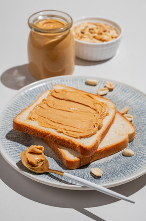 Peanut butter toast on a plate on a blue background with jar of spread, nuts, spoon and shadow. Concept of nutritious and healthy snacks for an American breakfast. Front view, copy space.の写真素材