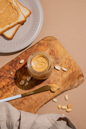 Peanut butter in a glass jar and knife on a wooden board on a beige background with toast, scattered...