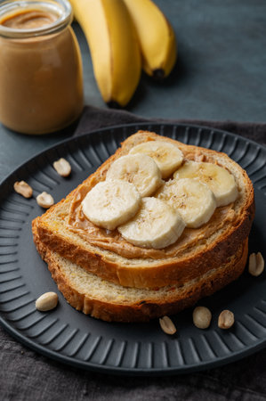 Peanut butter toast and banana on a black plate on a dark background with nuts, jar of spread and fruits close up. Concept for nutritious and healthy snacks for an American breakfast. Front view.の写真素材