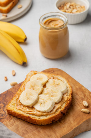 Peanut butter toast and banana on a wooden board on a light background with nuts and jar of spread. Concept for nutritious and healthy snacks for an American breakfast. Front view, copy space.の写真素材