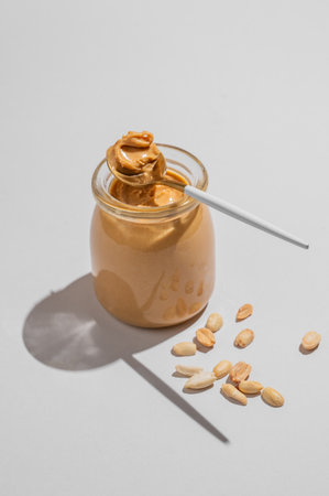 Peanut butter in glass jar on gray background with spoon, scattered salted nuts and shadow. Concept of tasty and healthy snack for sweet sandwiches for breakfast. Front view and copy space.の写真素材
