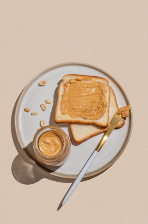 Peanut butter toast with jar of spread, nuts and knife on a plate on a light background. Concept of nutritious and healthy snacks for an American breakfast. Top view,  copy space.の写真素材