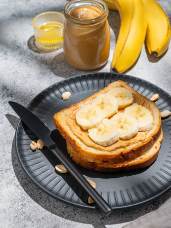 Peanut butter toast and banana on a black plate on a gray background with nuts, knife, fruits and morning shadow close up. Concept of sweet diet and healthy snacks for breakfast.の写真素材