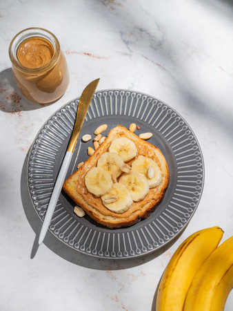 Peanut butter toast and banana on a dark plate on a marble background with fruits, knife and morning shadow. Concept of sweet diet and healthy snacks for breakfast. Top view, copy space.の写真素材