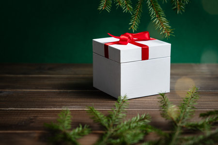 A white gift box with a red ribbon sits on a wooden table on a dark green background with fir branches and twinkling lights. A festive banner concept for a Christmas ad or card. Front view, copy space.の写真素材