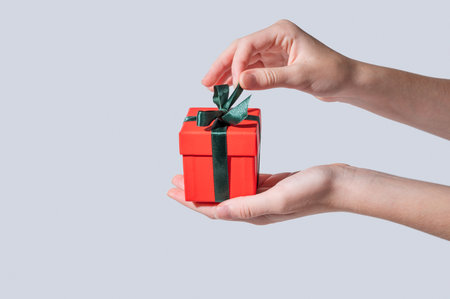 A hands opening  a small red gift box with an emerald ribbon on a light isolated background in a minimalist style. Concept advertising layout for a greeting card. Front view, copy space.の写真素材