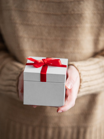 A close-up of a woman holding a white gift box with a red ribbon on a warm beige sweater with soft focus. Concept of holiday and home Christmas coziness. Front view.の写真素材