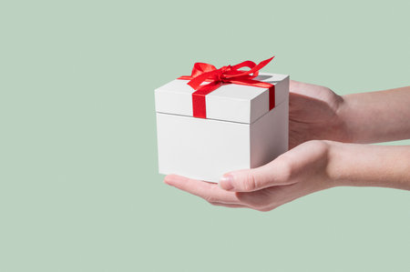 A hands holding a white gift box with a red ribbon on an isolated light green background in a minimalist style. Concept advertising layout for a greeting card. Front view, copy space.の写真素材