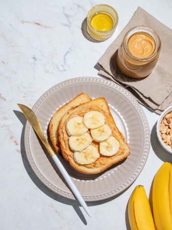 Peanut butter toast and banana on a plate on a marble background with fruits, nuts, honey and morning shadow. Concept of sweet diet and healthy snacks for american breakfast. Top view, copy space.の写真素材