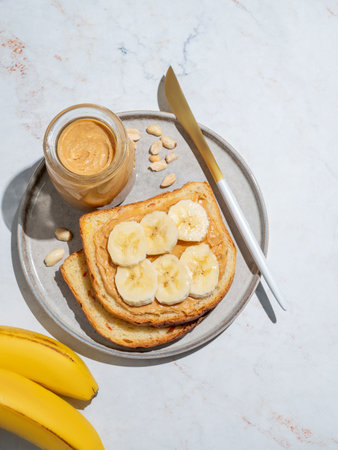 Peanut butter toast and banana on a plate on a marble background with fruits, nuts, knife and morning shadow. Concept of sweet diet and healthy snacks for american breakfast. Top view, copy space.の写真素材