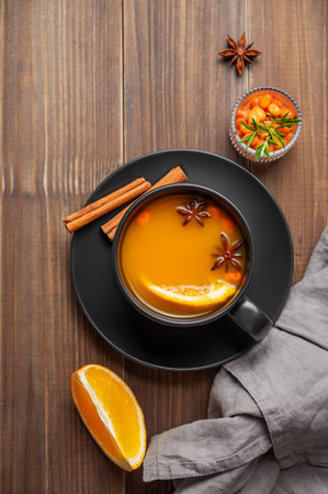 Sea buckthorn herbal tea in a black cup on a wooden background with spices, berries and napkin. Concept for a hot autumn organic drink to boost immunity in rustic style. Front view, copy space.の写真素材