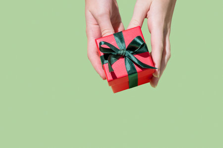 A hand holding a small red gift box with an emerald ribbon on an isolated pastel green background in a minimalist style. Concept advertising layout for a greeting card. Top view, copy space.の写真素材