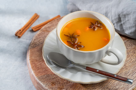 Sea buckthorn herbal tea in a cup on a wooden board on a light background with spices, napkin and morning shadows close up. Concept for a hot autumn organic drink to boost immunity. Top view.の写真素材