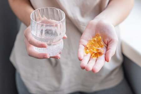 A close-up of a person's hands holding yellow softgels in one palm and a glass of water in the other. Concept of health, vitamins, or dietary supplements.の写真素材