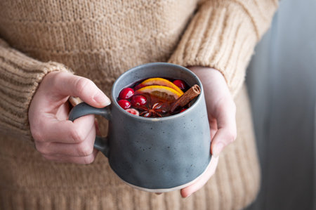 A woman holds a mug of non-alcoholic mulled wine or a spicy hot drink with orange, cinnamon, and cranberry on a beige sweater background close up. A homey winter concept. Copy space.の写真素材