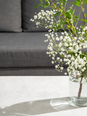 Glass vase with bouquet of gypsophila on light table and morning sunlight on grey sofa background....