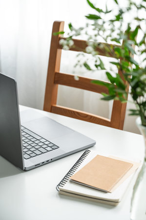 A laptop, notepad, and vase of flowers on a white table in natural light, against a wooden chair and...