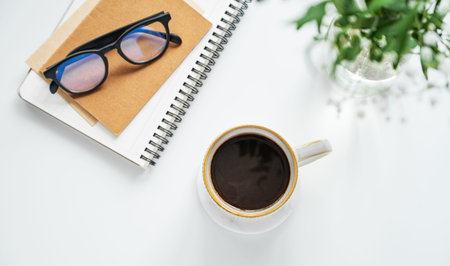 A cup of coffee, notepads, glasses, and a vase of flowers on a white table. Concept for a cozy and...