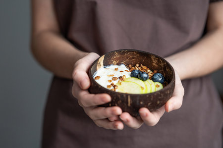 Bowl of granola with greek yogurt, apple and blueberries in hands of woman in brown t-shirt close-up. Milk breakfast concept, oat muesli in natural light. Front view, copy space.の写真素材