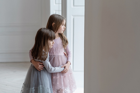 Serious little sisters in beautiful dresses stand near a window.の写真素材