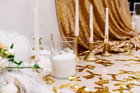 Bright golden foil and white candles in candlesticks on a bright parquet near the wedding table. Decoration of wedding in royal styleの写真素材