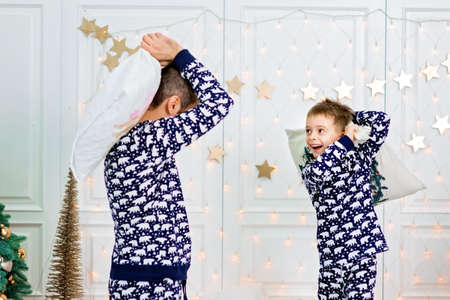 Happy family in pajamas having pillow fight on the bed indoors in light interior room near decorated Christmas tree. Scandinavian style. Togetherness conceptの写真素材